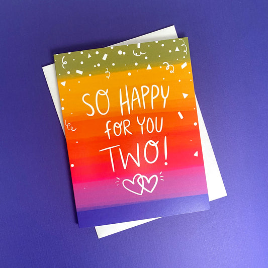 A greeting card with a colorful rainbow background with the words "So happy for you two" on the front. black inside size a2 greeting card with white envelope.